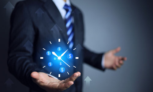 A man in a suit and tie holding a clock with dollar signs instead of numbers on the dial.